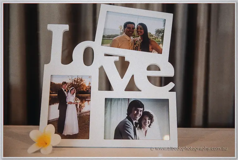 A white Love-letter frame holds family wedding portraits, a frangipani resting against it.