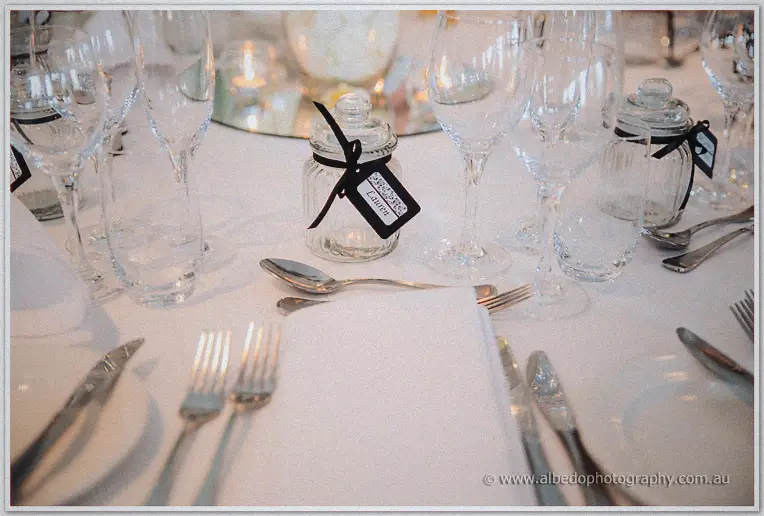 Close-up of a reception place setting with wine glasses, folded napkin and a ribbon-tied gift jar.