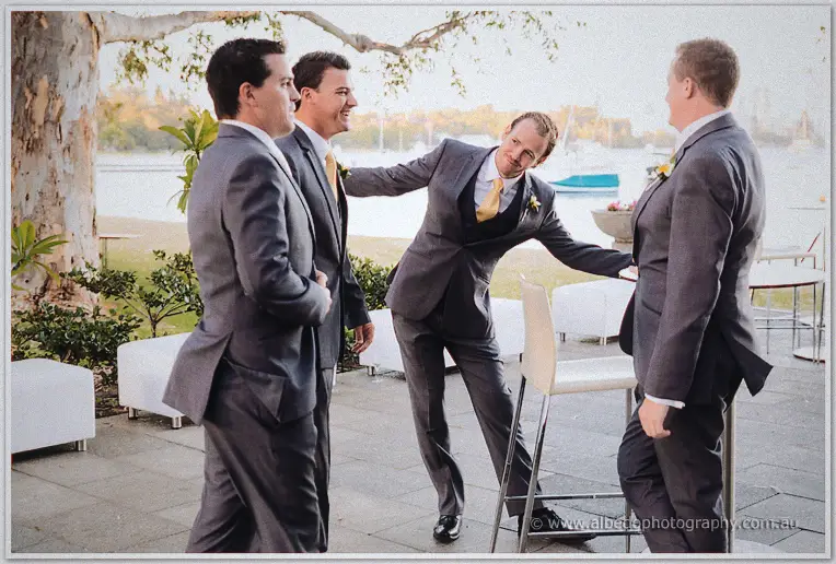 Groomsmen in grey suits and gold ties clown for the camera on a riverside terrace at dusk.