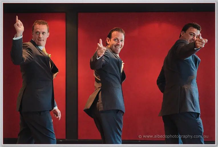 Three grinning groomsmen point finger-guns at the camera in front of a red-lit reception wall.