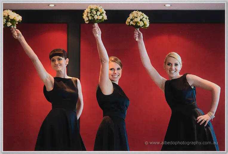 Bridesmaids in little black dresses raise their cream bouquets triumphantly against a red wall.