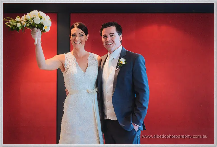 The bride thrusts her bouquet skyward as her pocketed-hand groom grins before the red wall.