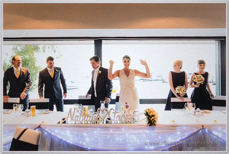 Bridal party enters the reception as the bride throws both arms up, fairy-lit table behind.