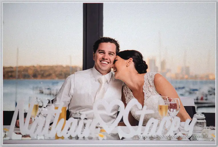 Seated at the bridal table, the couple nuzzle cheeks as the marina glows amber behind them.
