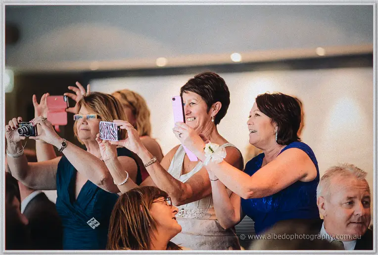 Guests crane forward with phones and cameras raised high, grinning as they capture the moment.