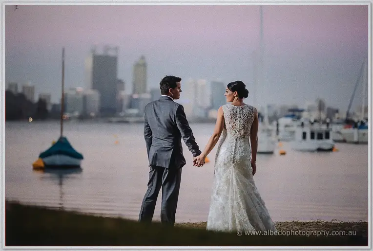 Hand in hand the couple face the river at twilight, moored boats and Perth towers in pink haze.