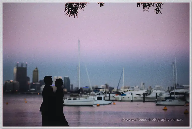 Silhouetted against a rose-pink sky, the couple embrace at the water's edge, masts soft behind.