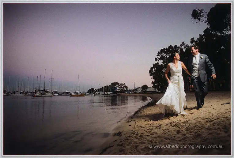 On the sandy Matilda Bay shore at dusk, the bride lifts her gown and strides the waterline with her groom.