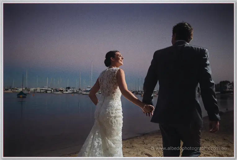 From behind, the bride beams up at her groom as they walk the beach, her lace train trailing.