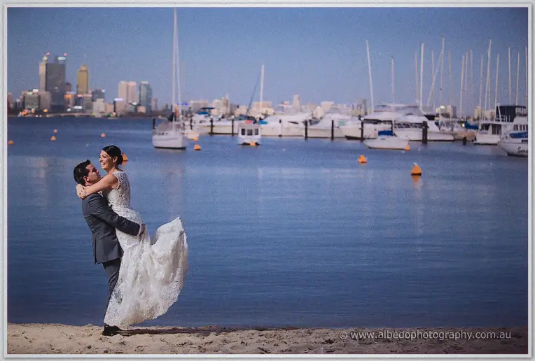Nathain lifting Stacey in his arms on the sand, Perth skyline and yacht masts glowing over Matilda Bay at dusk