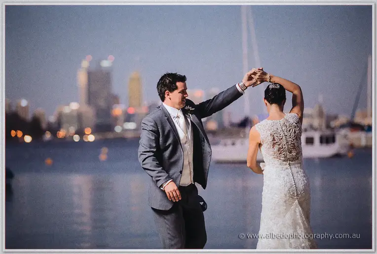 The groom twirls his bride by the river at nightfall, city lights sparkling across the water.