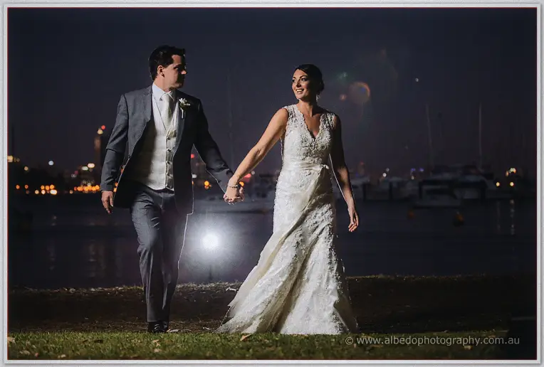 Hand in hand on the darkened lawn, the couple smile at each other under lamplight, city far behind.