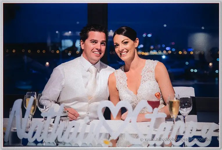 Nathain and Stacey beam from the head table at night, harbour lights twinkling through the window.