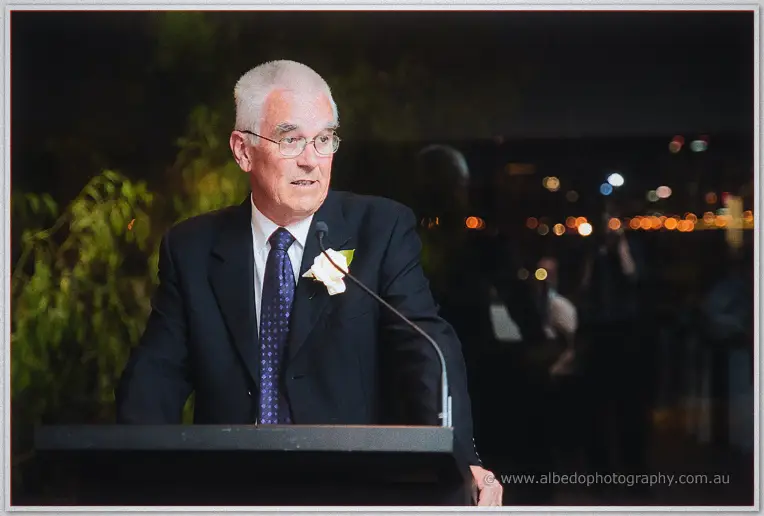 A white-haired father in a navy suit with a cream rose boutonniere gives his speech at the lectern.