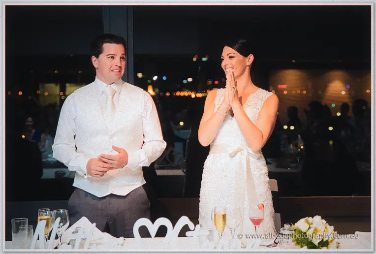 The bride presses her hands together laughing as the groom speaks, harbour lights glowing behind.
