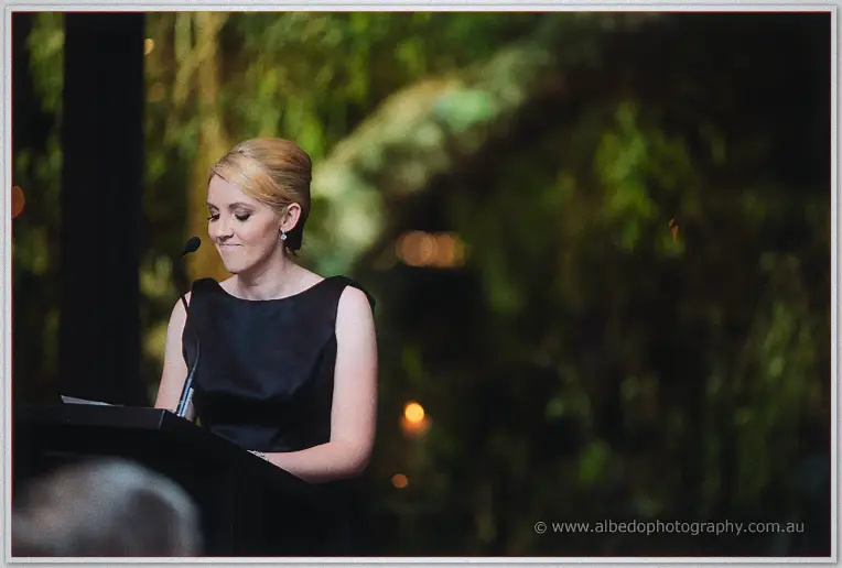 A blonde bridesmaid in a little black dress smiles down at her notes while speaking at the podium.