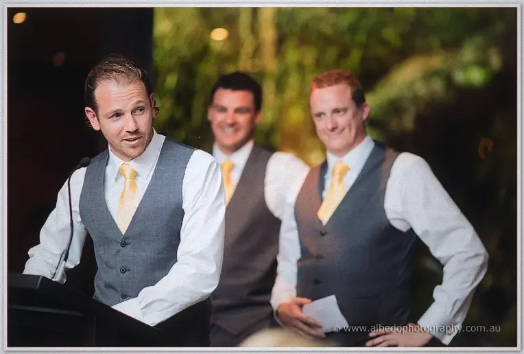 A groomsman in a grey vest and gold tie takes the mic as two grinning mates in vests crowd behind.