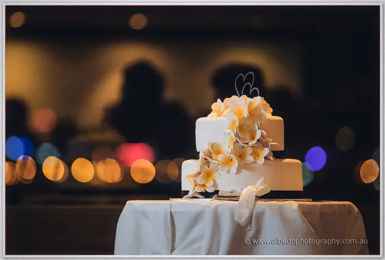 Two-tier white cake crowned with frangipanis and a wire heart, glowing against blurred night lights.