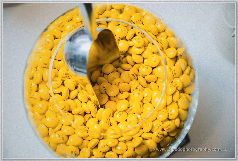 A glass bowl of yellow chocolate buttons with a silver scoop, close-up from the wedding lolly buffet.