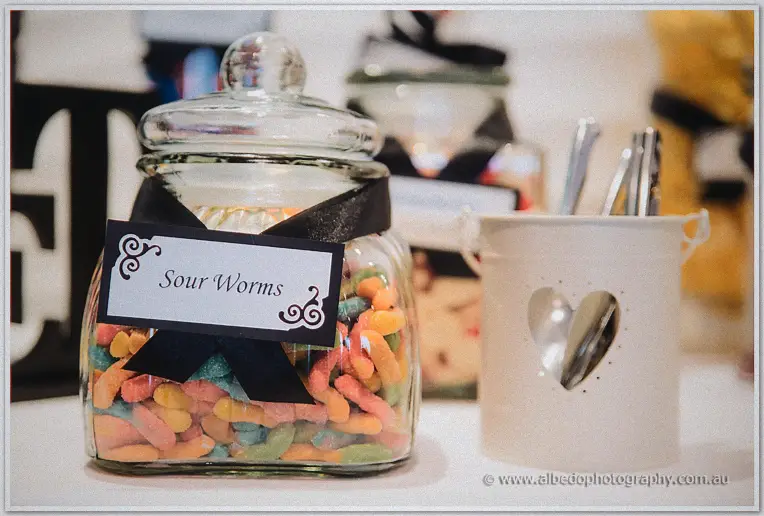A black-ribboned jar labelled Sour Worms sits beside heart-cutlery pots on the reception sweets table.
