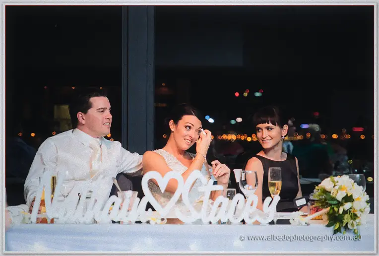 Dabbing a happy tear, Stacey laughs as Nathain's hand rests on her back, a bridesmaid smiling.