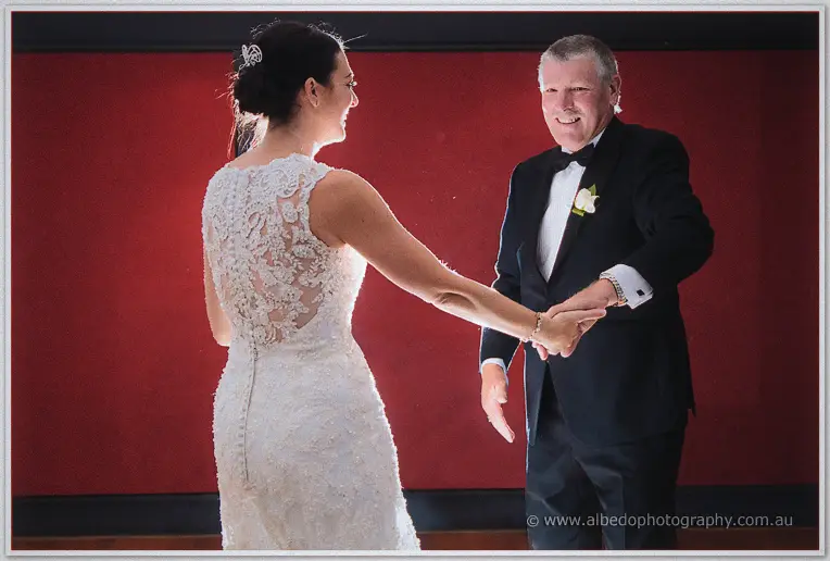 Stacey takes her smiling father's hand for a dance before a deep red backdrop.