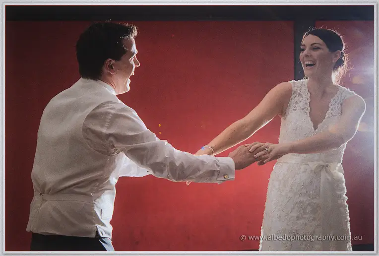 Nathain and Stacey laugh as they dance hand in hand against the red reception wall.