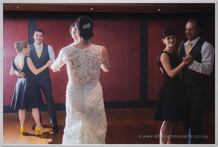 Stacey, seen from behind, dances with guests in dark dresses and yellow ties at the reception.