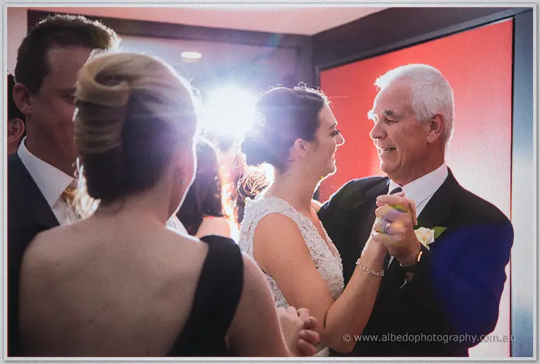 Stacey laughs in a lens flare while dancing with an older gentleman amid fellow guests.