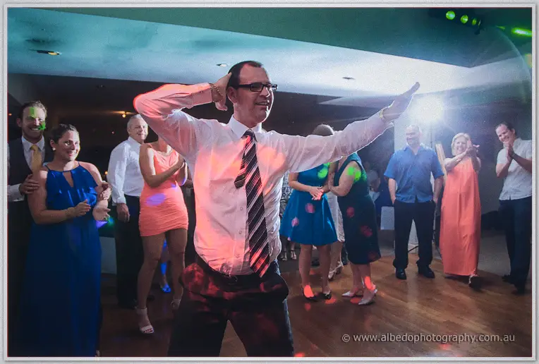 A guest in a striped tie strikes a dance pose on the floor as friends clap around him.