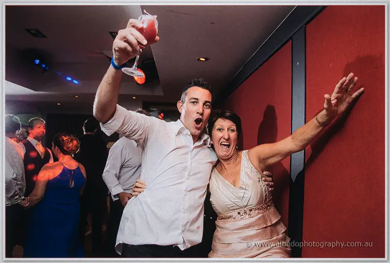 Two wedding guests cheer with a raised cocktail by the red wall during the party.