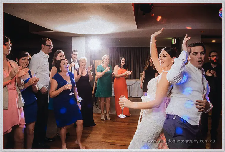 Stacey and Nathain dance back to back, champagne in hand, while guests clap along.