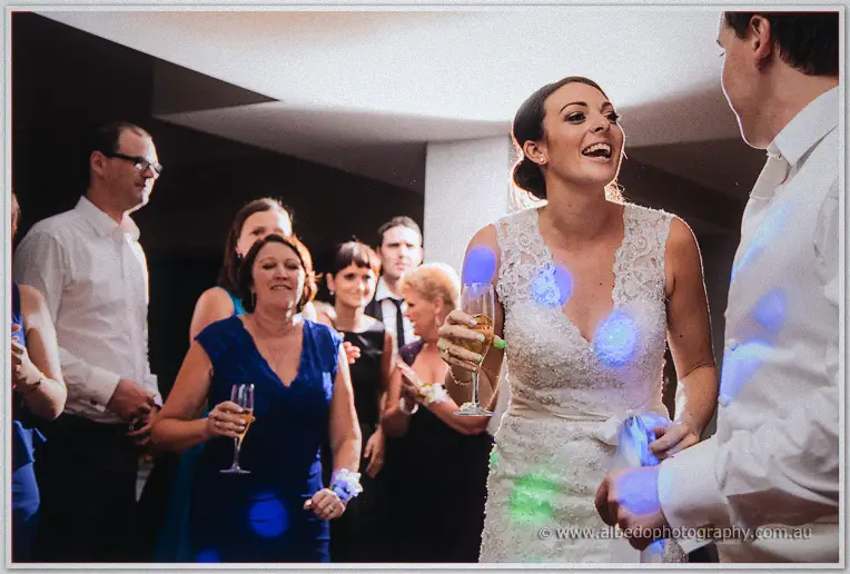 Stacey grins mid-dance, champagne flute in hand, under blue disco dots among clapping guests.