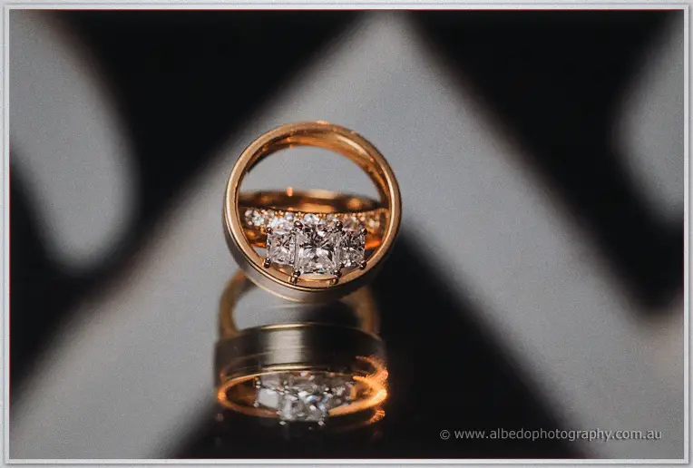 A diamond trilogy ring and gold bands rest mirrored on a dark reflective surface.