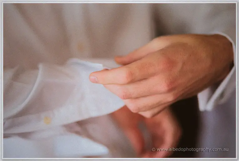 Close-up of Mitch's fingers fastening a cufflink on his crisp white dress shirt in soft morning light