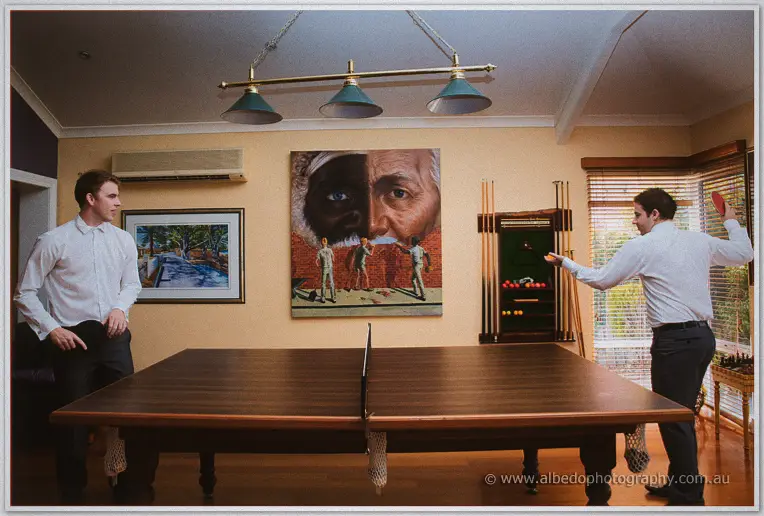 Two groomsmen in white shirts play table tennis across a timber table in the warm-lit games room