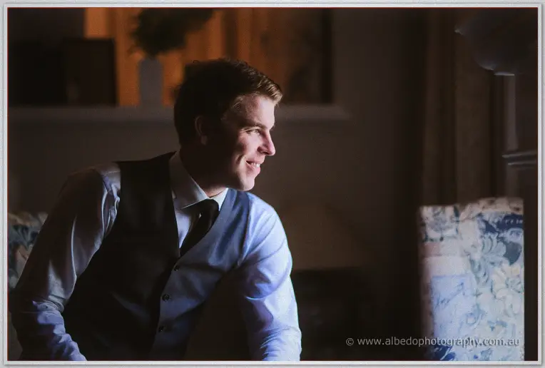 Mitch smiles in his grey waistcoat and tie, soft lamp light warming the room as he waits