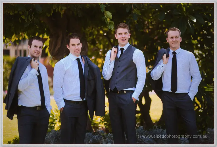 Groomsmen stand shoulder to shoulder in the shade, jackets slung over shoulders and ties in place