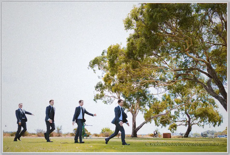 Groomsmen strike a playful synchronised strut across the park lawn under a big eucalypt