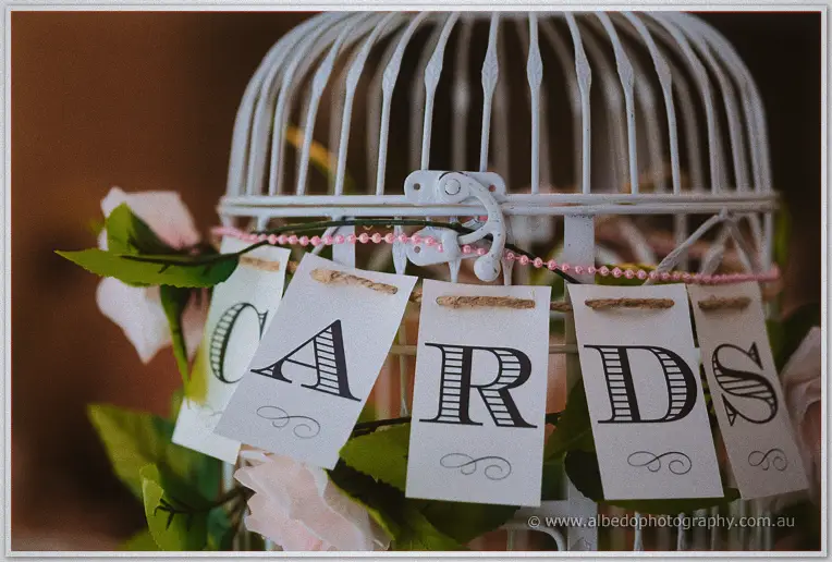 A white birdcage strung with pink beads displays hanging cards spelling CARDS among fabric roses