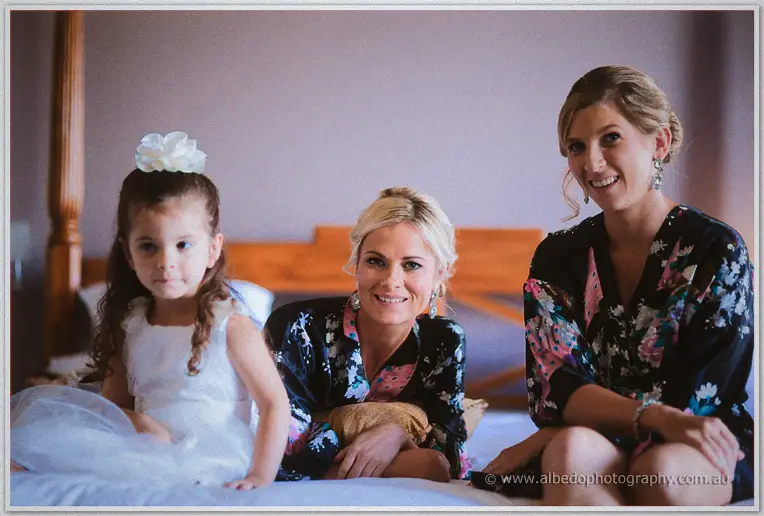 Flower girl in white sits on the bed with two bridesmaids in matching floral robes