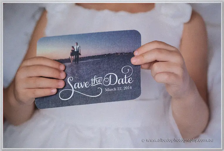 Small hands in a white dress hold a navy save the date card showing Cristina and Mitch on the beach at dusk