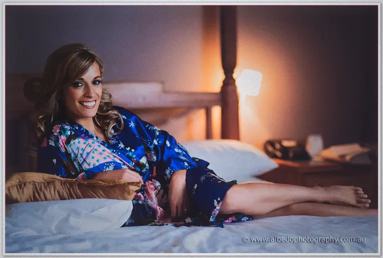 Cristina lounges across the hotel bed in her blue robe, warm lamp glow behind her