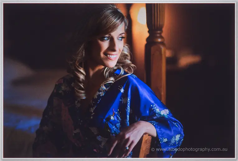 Cristina smiles over her shoulder from the bedpost, lamplight glowing on her blue robe