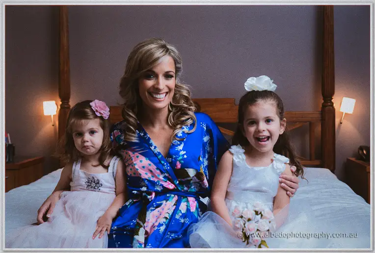 Cristina beams between two flower girls in white on the bed in the warm bedroom