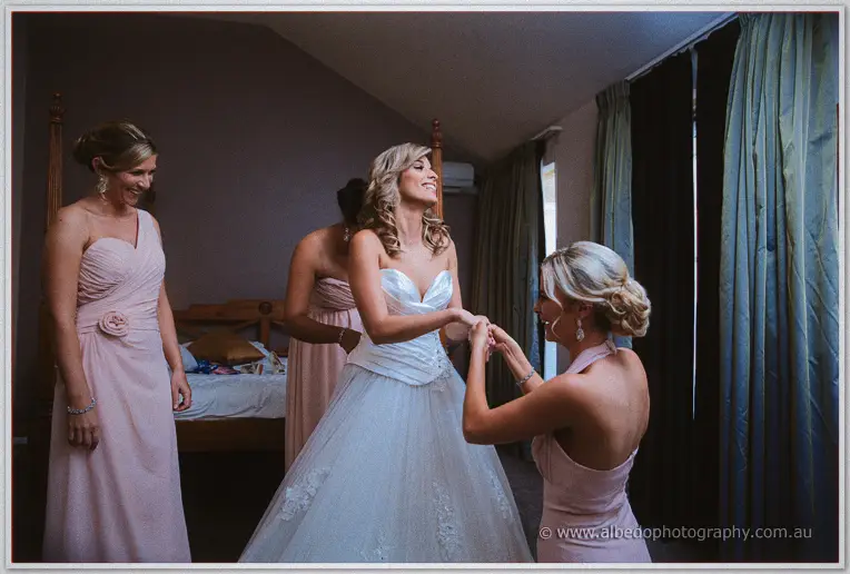 Bridesmaids in blush pink lace up Cristina's gown as she laughs in the bedroom