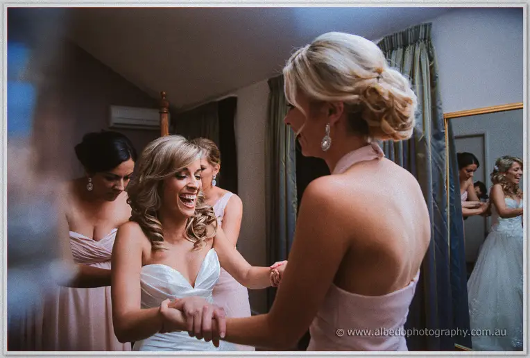 Cristina laughs as her bridesmaids clasp her bracelet in the bedroom