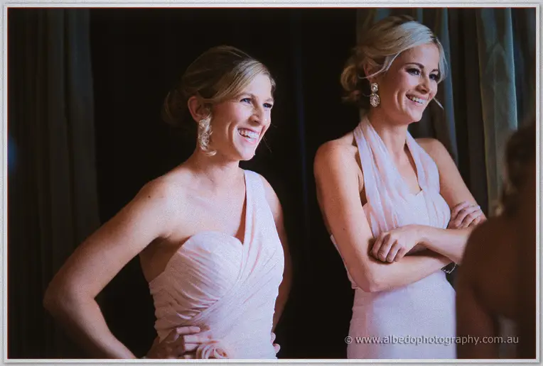 Two bridesmaids in blush gowns grin as they watch Cristina get ready