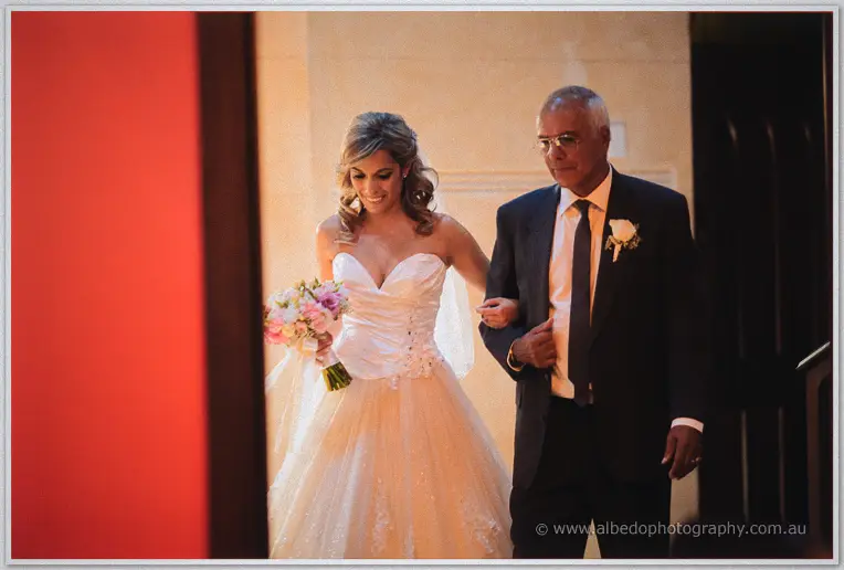 Cristina arrives on her father's arm, pink bouquet in hand, framed by warm light at the basilica doorway