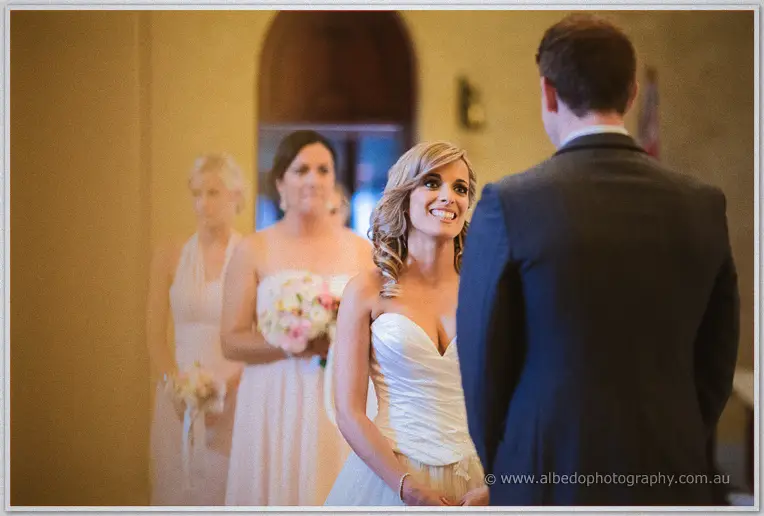 Cristina beams up at Mitch as blush-toned bridesmaids look on during the ceremony at St Patrick's